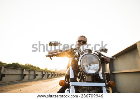 Brutal portrait handsome biker man in sunglasses sits on a bike on a sunset on old rusty metal bridge in city against house and sunny sky background Empty copy space for inscription