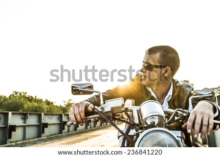 Romantic portrait handsome biker man in sunglasses sits on a bike on a sunset on old rusty metal bridge in city against house and sunny sky background Empty copy space for inscription