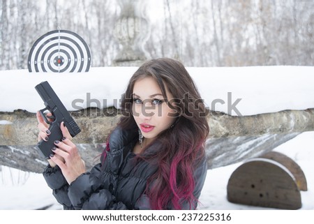 Portrait of young woman with snowflake in red brunette curly hair Cute girl with black pistol gun against  snowdrift cold winter trees in city park on old retro vintage fountain in forest background