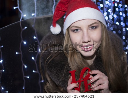 outdoor outside portrait of happy young Christmas woman with Santa Claus red hat Blue bokeh blur light effect. Cute girl with smiley face looking at camera Female holding word love Night light