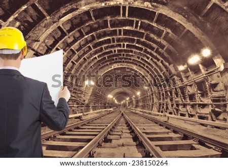 engineer hand wear yellow helmet for workers security holding empty blue print paper document against  background of an underground mine with arc legs and rails for trolleys with coal in perspective