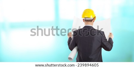 Rear view of architect looking comparing blue print paper plan housing project with building wearing yellow helmet Back view businessman on wall and  window background Empty copy space for inscription
