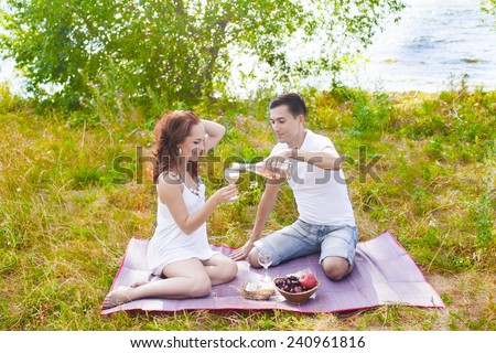 couple celebrating at picnic Man pour champagne woman cake in plate girl wearing summer white very sexy hot short dress They looking at wine glass against green grass field, blue sea water texture