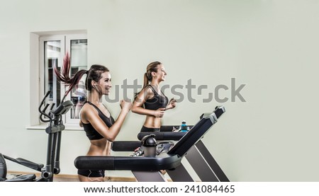 Portrait of Two young adult cute sporty women run on machine in the gym centre Beautiful girls with white headphone inside on on wall and window background Couple of  sport girls with fly hair