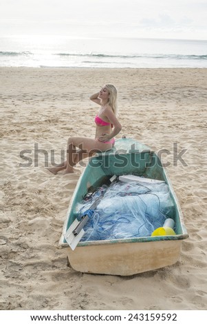 Beautiful sexy blond girl in pink bathing suit sitting alone on wooden plastic Cute woman near old boat on sand tropical beach against water texture and blue sky with sun set on background