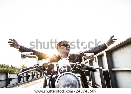 Brutal portrait biker man in sunglasses sit on chopper motor bike on sunset sky Stand old rusty metal bridge in city against house and sunny heaven background Empty copy space for inscription Hands up