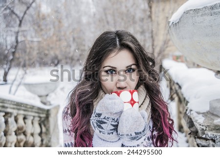 Portrait of Woman with cup in winter snowy park Young adult girl with wide eyes looking at camera against old retro vintage architecture concrete figure fence Snowflake in dark with red long hair tip