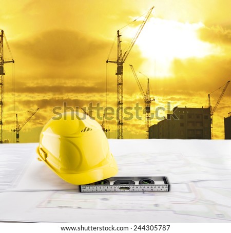 Objects of safety helmet level lie blue print architect paper plan on table with sunset sky with Nature scene building construction crane lift load against evening sun rise shine and new modern house