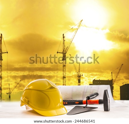 Mason tools yellow  helmet hammer trowel lie blue print architect paper plan on table with sunset sky with building construction crane lift load against evening sun rise shine and new modern house