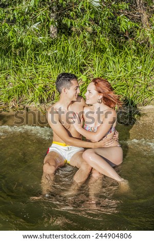 young adult embracing couple in love at sand tropical beach background Copy space for inscription Beautiful man and woman sit in clear blue water with wave against green fresh grass trees in jungle
