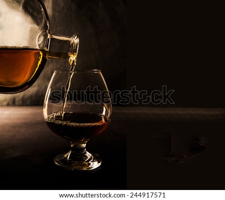 Bottle pour wineglass with scotch or cognac on dark background at night time Transparent glass stand on table on black wall texture background Empty space for inscription