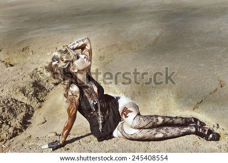 Portrait of beauty freak tattoo girl with bat and with body art on her hand on empty land desert background woman paint body art in form of fire in shirt and jeans shorts and torn raggy pantyhose