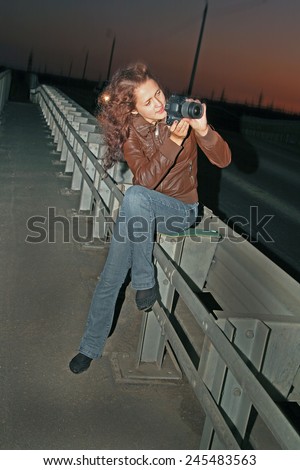 Cute girl full length tourist sit on metal car bridge  taking photos with black modern photo camera Woman wear casual dress blue sexy jeans on slim long legs and brown leather jacket Night image