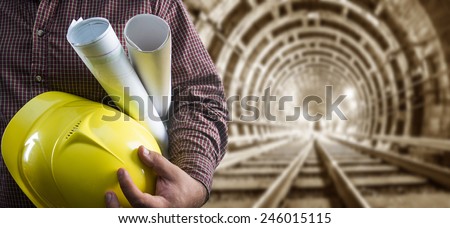 engineer hand holding yellow helmet for workers security and blueprint paper plan against the background of an underground mine with arc legs and rails for trolleys with coal in perspective