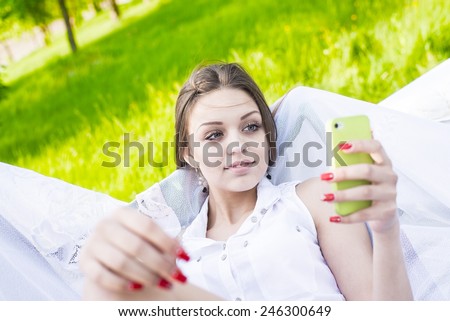 Cute slim girl looking at cell screen read book or watch video on summer fresh green grass background Portrait of Beautiful young adult caucasian woman reading a message on mobile phone