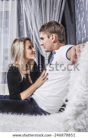 Portrait of funny Young adult romantic couple sitting on white wool sofa inside room against white transparent curtains window Man and girl look each other pull a face ape, fooling around