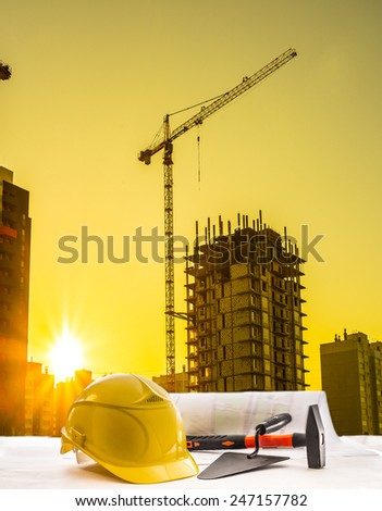 Mason tools yellow helmet hammer trowel lie blue print architect paper plan on table with sunset sky with building construction crane lift load against evening sun rise shine and new modern house
