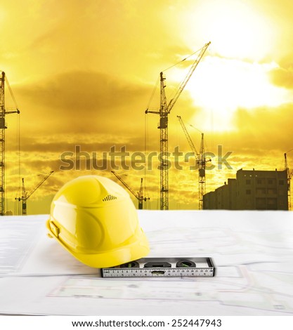 Objects of safety helmet level lie blue print architect paper plan on table with sunset sky with Nature scene building construction crane lift load against evening sun rise shine and new modern house