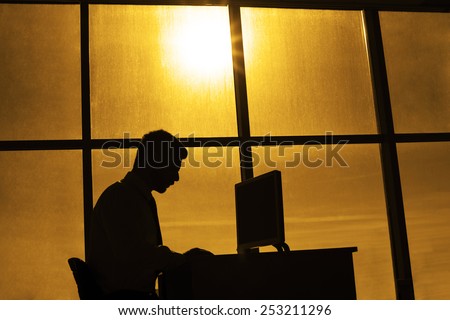 silhouette of business man on  background office room window of beautiful sunset sky with sun shine rays alone Businessman sit and type print text on pc computer monitor screen Idea late hard work