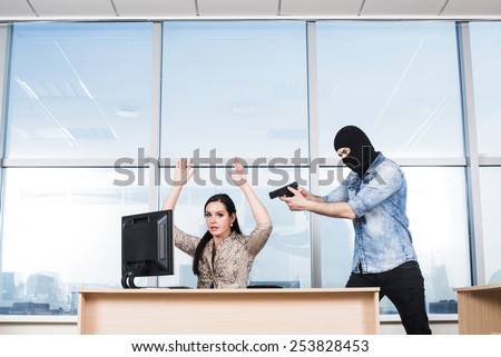 Portrait of man wearing black balaclava with gun looking at hostage scared business woman on blue sky window background Empty space for inscription Girl sit on table near pc computer Hands up