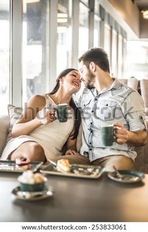 Happy couple enjoying a coffee at the coffee shop cafe Young adult man and woman kiss touch nose each other against window glass in perspective Romantic relationship Pair hold green mug in hands