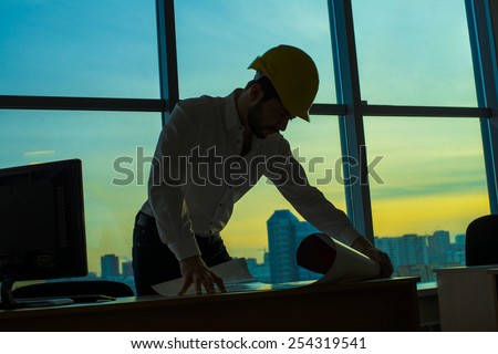 Silhouette builder engineer wear security helmet look at blueprint paper construction drawing plan on  background of sunset window frame blue yellow sky with clouds near pc monitor computer