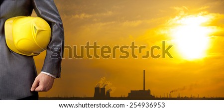 Hand or arm of engineer hold yellow plastic helmet for workers security over Central Electric Heat CHP in winter Empty space for inscription Yellow sunset sky with clouds
