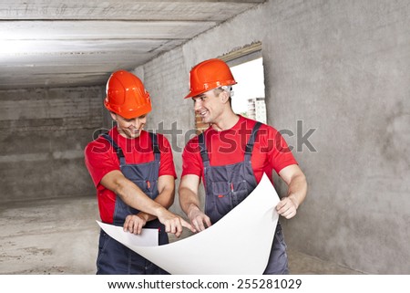 two builder engineer Architects working in empty concrete room  - planning and looking at blueprint hold in hands  Builder wear blue uniform and red hardhat shirt overall Man show in paper plan