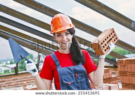 portrait of female woman mason hold in hand red brick with hole metal trowel against metal beam roof and blue summer sky with clouds Cute girl wear red plastic helmet shirt and blue overall uniform