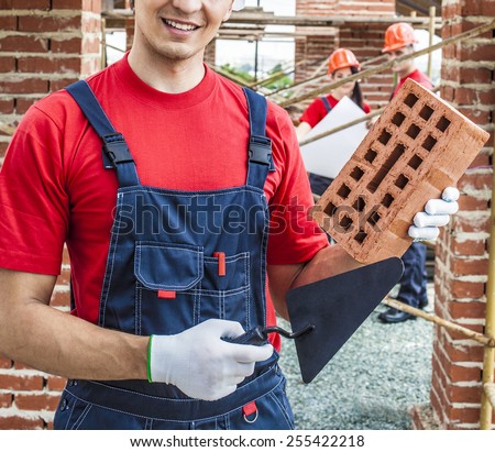construction mason worker bricklayer hold in hand red brick with trowel putty knife outdoors wear white gloves and blue cotton material overall against two engineer with blueprint paper plan
