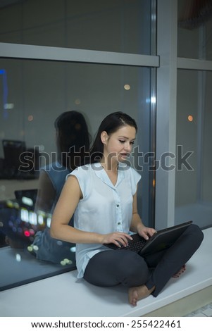 full length portrait of Romantic Girl With laptop stand on long legs lie along dark night window View on city town with bokeh light Reflection in glass Hands on knee Female cute woman look at screen