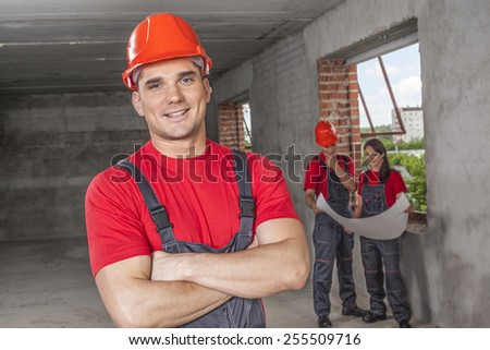 Portrait of cheerful handsome young adult  industrial engineer wear plastic helmet uniform overall and red cotton shirt look at camera against two worker with blueprint paper plan near window frame