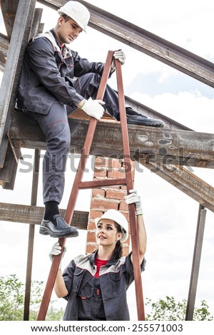 Full length portrait of worker man on steel beam raises his hands sitting on rusty metal beam - roof frame, Under construction on blue summer sky background with clouds woman wear white plastic helmet
