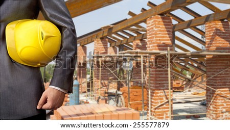 torso of engineer worker holding in hands yellow plastic helmet hardhat for workers security on background of  new metal red brick roof and blue summer sky with clouds Empty space for inscription