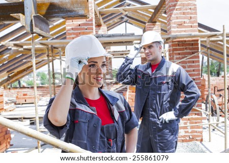 working couple - woman and man - two Young adult builders on a background of iron metal beam roof trusses and summer city town with green trees at street , blue sky with clouds on backdrop