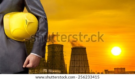 Hand or arm of engineer hold yellow plastic helmet for workers security over Central Electric Heat CHP in winter Empty space for inscription Yellow sunset sky with clouds