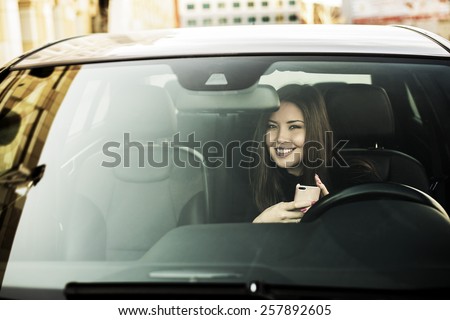 Portrait of beautiful asian brunette young woman holding in hands cell mobile phone inside new car through transparent window background Copy space for inscription Taxi Girl call customer Empty place