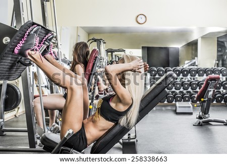 Portrait of two young Girls do exercise for legs and hands. in fitness gym on mirror with reflection and window background 2 woman with long blond and brown hair sitting No face Unrecognizable person
