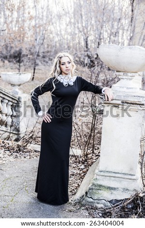 tall beautiful lady in long black retro dress standing on dry branches based on old abandoned fence on brown building wall with windows and looking at far away Girl with long blond curly hair
