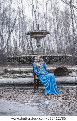 Full length portrait of Beautiful woman sit on wooden old retro vintage aged chair Lady wear blue long elegant dress Brown Fallen leaves lie on land Autumn cold season against park trees and gray sky