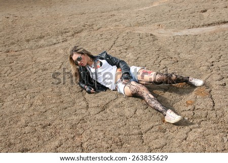 Full length Portrait of stylish tattoo woman painted body art in the form of red blue flower in the white t-shirt, black leather jacket and denim shorts and torn pantyhose on background of gray desert