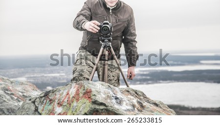 No face Unrecognizable person Professional photographer shooting Finger push button Man stand on stone mountain high in heaven against horizon with town and river or lake Photo Camera stand on tripod