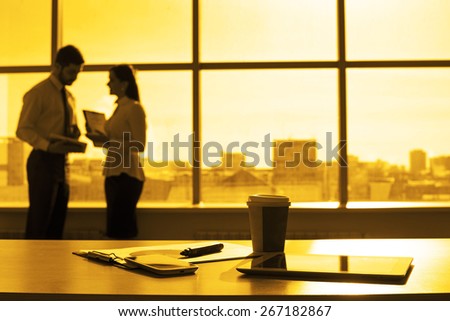 Business tools in office room tablet computer paper cup with hot coffee Cell mobile phone Paper lie on wooden table against blue sky window with houses Man and woman stand with document in hand