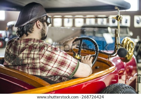 man looking at auto through old retro vintage aged  car window Hand lie on wheel  Beard Gay wear  protective glasses from leather and glass goggles and cap Near door weighs spare wheel