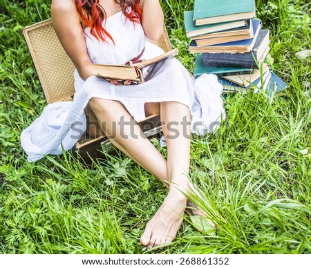 No face girl in spring blooming park Hand holding old retro open book Woman sitting in vintage aged suitcase on green grass against fresh green grass background near stack