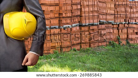 torso of engineer worker holding in hands yellow plastic helmet hardhat for workers security on background of new red brick pile on pallet and green summer grass Empty space for inscription