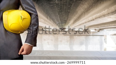 worker or engineer holding in hands yellow helmet for workers security on background of  new concrete bridge and metal tube over river in perspective buildings gray idea building new road junctions