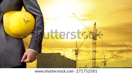 Silhouette Crane lifts load engineer hold yellow helmet for workers security on background of new highrise apartment buildings and construction cranes on backdrop of evening sunset cloudy sky