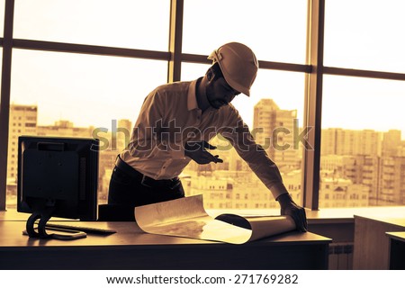 Silhouette builder engineer wear security helmet look at blueprint paper construction drawing plan on background of sunset window frame blue yellow sky with clouds near pc monitor computer