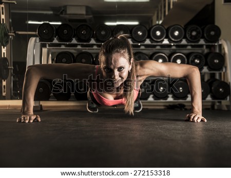 Young girl do push ups on dusk floor View of sexy cute sporty woman with strong hand Female look at camera with smiley face against dumbbell shelf mirror reflection on wall in gym studio room space